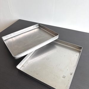 Puede incluir: Dos bandejas de horno rectangulares de metal con bordes elevados. Las bandejas están hechas de un material de color plateado, liso y ligeramente reflectante. Una bandeja está colocada parcialmente sobre la otra, mostrando una base plana y lados verticales.