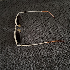 May include: Aviator-style sunglasses with a silver frame and dark lenses. The temples are a copper color. The sunglasses are lying on a dark, textured surface.