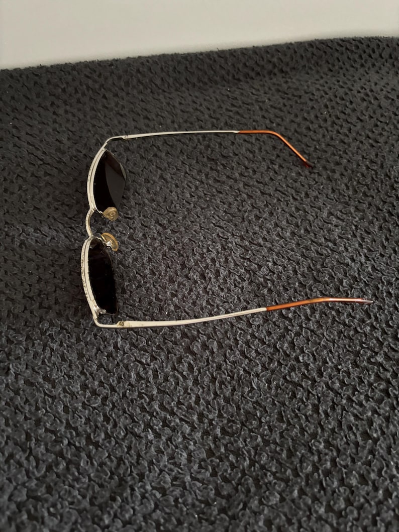May include: Aviator-style sunglasses with a silver frame and dark lenses. The temples are a copper color. The sunglasses are lying on a dark, textured surface.