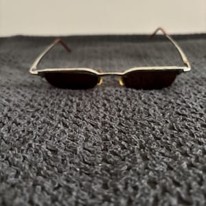 May include: A pair of rectangular sunglasses with a thin, silver-colored metal frame and dark lenses. The temples are a reddish-brown color. The sunglasses are resting on a textured, dark gray surface.