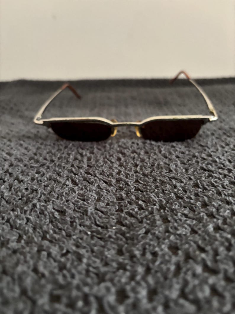 May include: A pair of rectangular sunglasses with a thin, silver-colored metal frame and dark lenses. The temples are a reddish-brown color. The sunglasses are resting on a textured, dark gray surface.