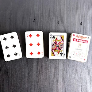 Puede incluir: Un juego de cartas que incluye un seis de picas, un seis de diamantes, una reina de tréboles y una caja con la etiqueta Amaro Montenegro Ti Regala Dal Negro. Las cartas están dispuestas en fila, cada una marcada con un número del uno al cuatro.