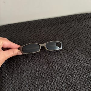 May include: Sleek, silver-framed sunglasses with dark lenses. The sunglasses have a rectangular shape and a modern design. The image shows the sunglasses against a dark, textured background.