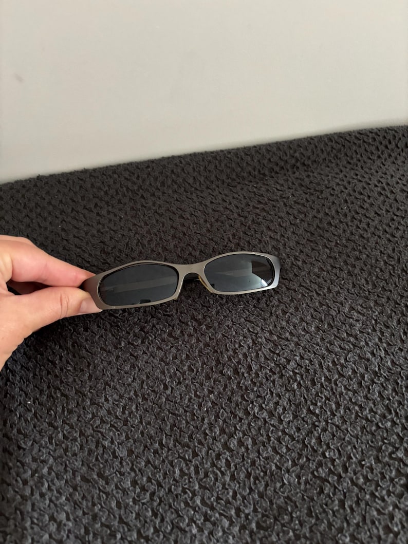 May include: Sleek, silver-framed sunglasses with dark lenses. The sunglasses have a rectangular shape and a modern design. The image shows the sunglasses against a dark, textured background.