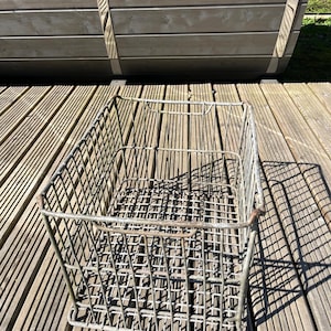 May include: A rectangular metal wire basket with a grid design, featuring a handle on each side. The basket is grey and rests on a wooden deck. A wooden structure is in the background.