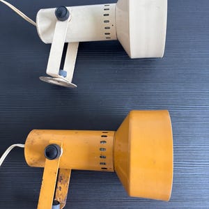 Könnte beinhalten: Zwei Metall-Strahler, einer in Off-White und einer in Senfgelb. Jede Lampe verfügt über einen zylindrischen Körper mit einem breiten, ausgestellten Kopf, eine Montagehalterung mit rundem Fuß und schwarze Einstellknöpfe an den Seiten des Gehäuses.