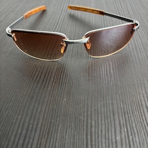 May include: Rimless sunglasses with brown tinted lenses and silver frames. The temples are a light brown color. The sunglasses are resting on a dark wood-grain surface.