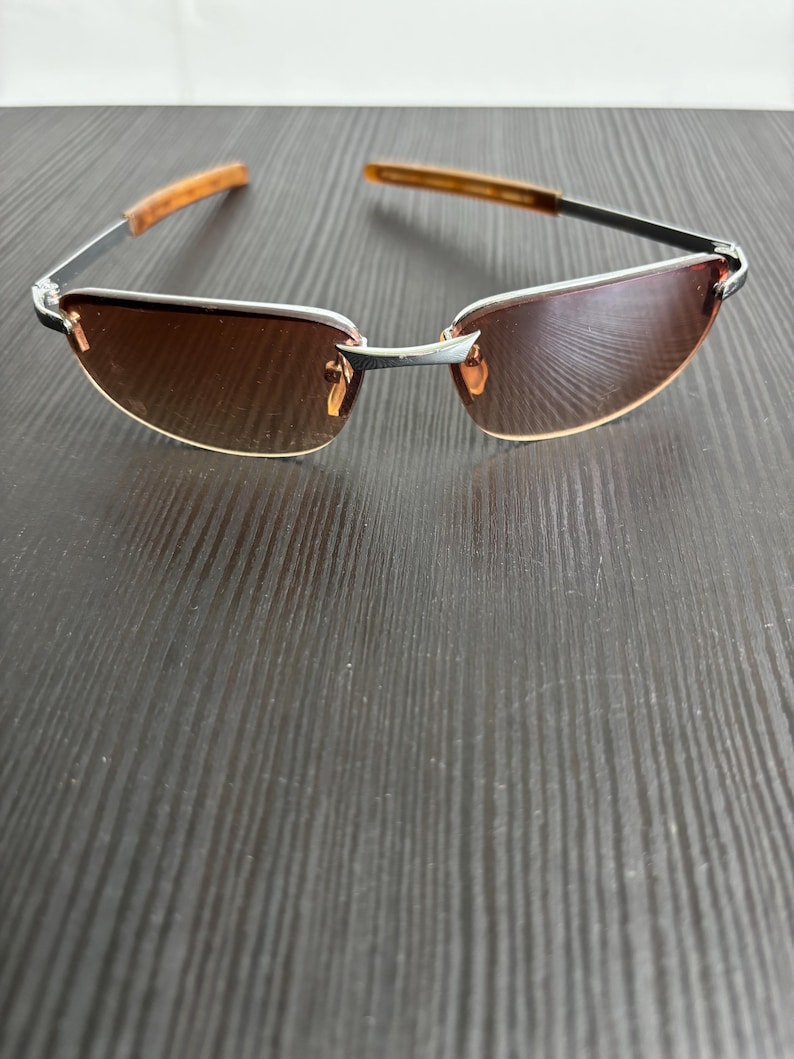 May include: Rimless sunglasses with brown tinted lenses and silver frames. The temples are a light brown color. The sunglasses are resting on a dark wood-grain surface.