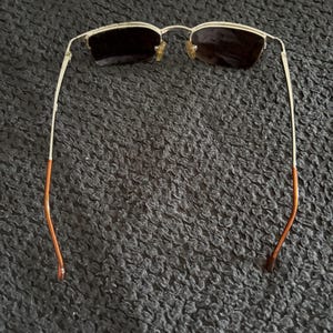 May include: Gold-framed sunglasses with dark lenses. The frames have a rectangular shape and a thin, delicate design. The temples are gold with orange tips. The sunglasses are set against a dark, textured background.