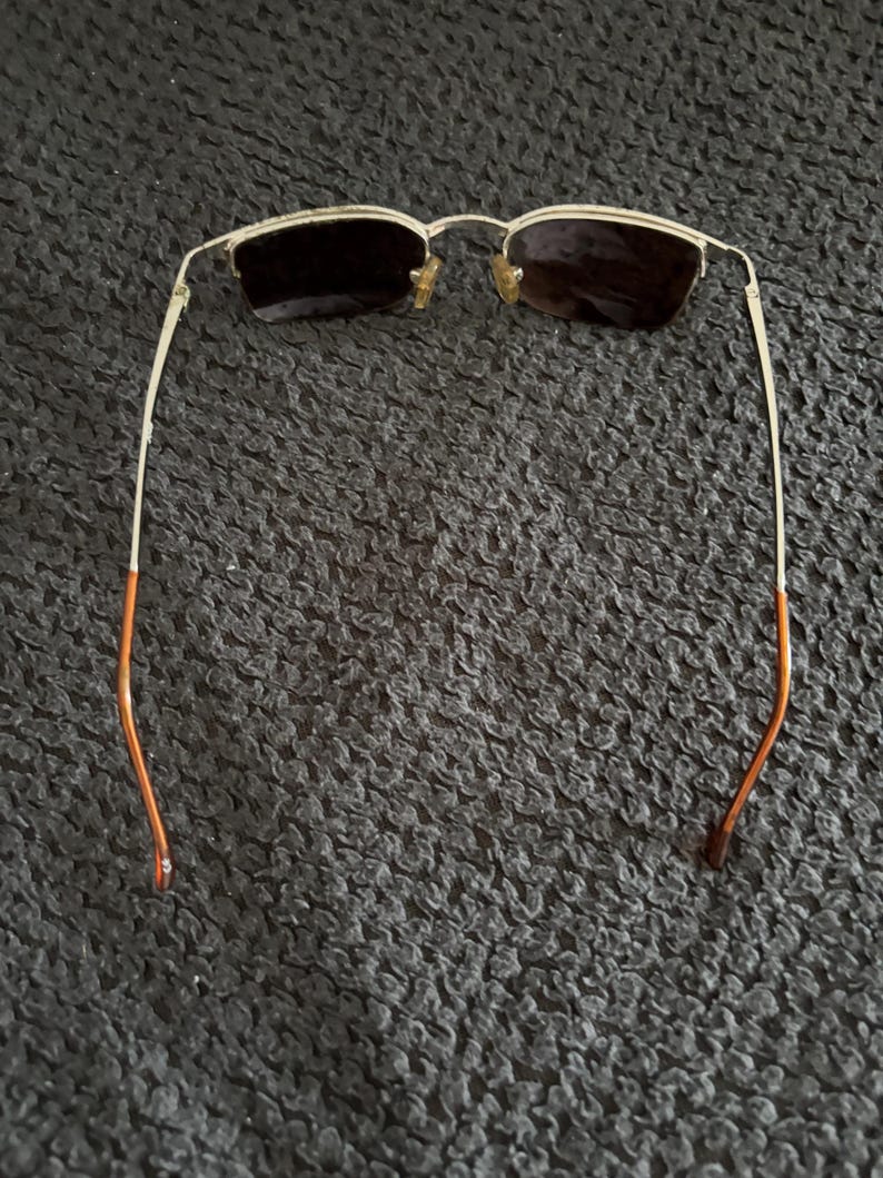 May include: Gold-framed sunglasses with dark lenses. The frames have a rectangular shape and a thin, delicate design. The temples are gold with orange tips. The sunglasses are set against a dark, textured background.