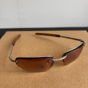 May include: Rimless sunglasses with brown tinted lenses and silver metal frames. The temples are a dark brown color. The sunglasses are resting on a brown surface.
