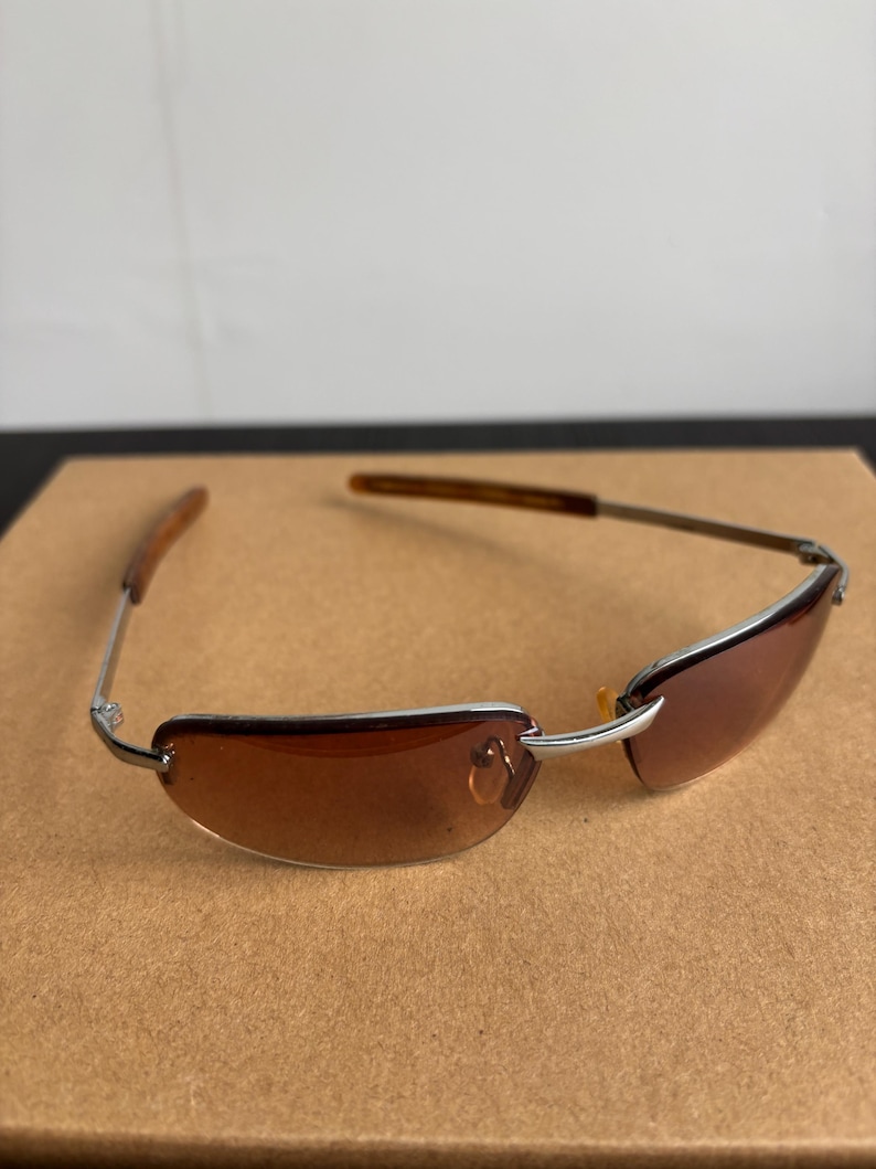 May include: Rimless sunglasses with brown tinted lenses and silver metal frames. The temples are a dark brown color. The sunglasses are resting on a brown surface.