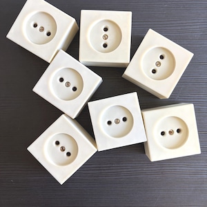 May include: A collection of six off-white square electrical wall outlets. Each outlet features a circular center with two pin holes and a central screw. The outlets are arranged in a scattered pattern, showcasing their uniform square shape and smooth finish.