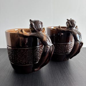 Puede incluir: Dos tazas de cerámica marrón con asas en forma de mono. Las tazas tienen una banda texturizada en el centro y un acabado brillante. Las asas de mono son detalladas y añaden un toque único a las tazas.