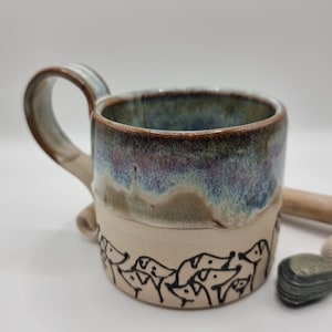 May include: Handmade ceramic mug with a unique design. The mug features a cream-coloured base adorned with a black dog illustration. The top rim has a gradient of blue, brown, and green. The handle is a matching brown colour.