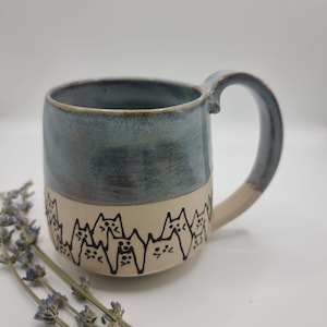 May include: A ceramic mug with a blue-grey glazed top and a cream base. The base has a black cat silhouette design. The mug has a curved handle. It is set against a white background with lavender sprigs.