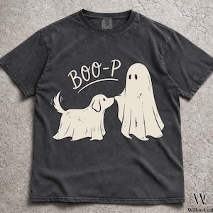 May include: A charcoal gray short-sleeve t-shirt featuring a cream-colored graphic of a dog wearing a ghost sheet and a standing ghost figure. The text BOO-P is printed in a curved font above the figures. The shirt has a crew neckline.