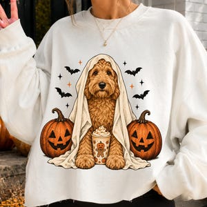 Puede incluir: Una sudadera blanca de manga larga con un gráfico de un perro de color marrón dorado envuelto en una sábana blanca como un fantasma, flanqueado por dos calabazas, murciélagos, estrellas y un vaso con un diseño de hombre de jengibre.