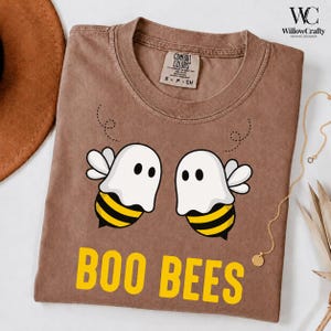 May include: A brown short-sleeve t-shirt featuring a graphic of two bees wearing white ghost costumes. The bees have yellow and black striped bodies and white wings. Below the graphic, the text BOO BEES is printed in bold yellow capital letters.