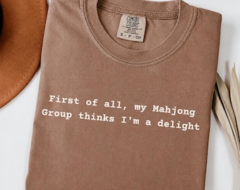 May include: A brown crew neck t-shirt with the white text "First of all, my Mahjong Group thinks I'm a delight" printed on the front. The shirt features a Comfort Colors brand label at the inner neckline, indicating a size small, made in Honduras.