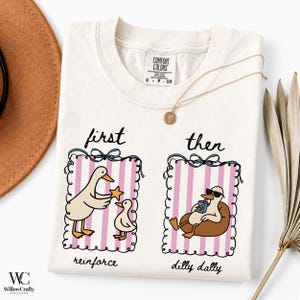 Puede incluir: Una camiseta de manga corta color crema con un gráfico de dos marcos a rayas rosas y blancas. El marco izquierdo muestra un pato dando una estrella a un patito con el texto first reinforce. El marco derecho muestra un pato con gafas de sol bebiendo de un cartón de zumo con el texto then dilly dally.