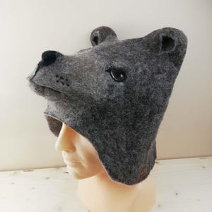 Könnte beinhalten: Eine dunkelgraue Filzwollmütze in Form eines Bärenkopfes mit abstehenden Ohren, einer Schnauze mit schwarzer Nase und dunklen Augen. Die Mütze bedeckt den Kopf und die Seiten, wobei Ohrenklappen nach unten reichen und das Gesicht einrahmen.