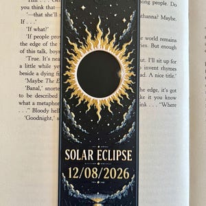 Puede incluir: Un marcapáginas rectangular negro con un gráfico de eclipse solar en dorado y blanco con rayos, rodeado de pequeñas estrellas y nubes. El texto "SOLAR ECLIPSE" y la fecha "12/08/2026" están impresos en letras doradas en la parte inferior.