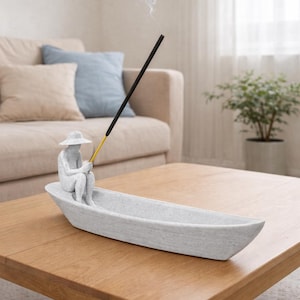 Könnte beinhalten: Ein grauer Räucherstäbchenhalter in Bootsform mit einer kleinen Figur einer Person mit Hut, die einen Räucherstäbchen hält. Der Räucherstäbchen ist schwarz mit einer goldenen Spitze, und Rauch steigt auf. Der Halter steht auf einem Holztisch.