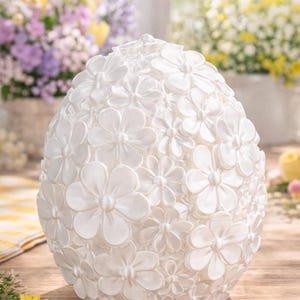 May include: A white, egg-shaped ornament adorned with a profusion of three-dimensional white flowers. The flowers, of varying sizes, are closely arranged, forming a textured exterior. It rests on a wooden surface, with floral arrangements visible in the background.