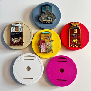 Op de afbeelding: Een verzameling kleurrijke ronde displaybases met decoratieve pins. De pins bevatten tekst zoals "Jurassic Coast", "Yellowstone National Park" en "Hadrian's Wall". De bases zijn in verschillende kleuren, waaronder wit, roze, geel, rood en blauw.