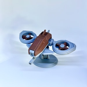 Puede incluir: Modelo impreso en 3D de una pequeña aeronave futurista. El modelo presenta un cuerpo central marrón con dos hélices azules y una base gris. El diseño sugiere un dron o nave espacial en miniatura, mostrando una mezcla de tecnología y diseño.