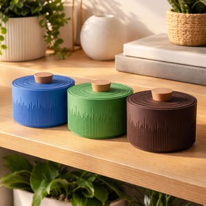 May include: Three round containers in blue, green, and brown, each with a wooden knob. The containers have a textured, vertical line design on the sides. They are displayed on a wooden shelf with plants in the background.