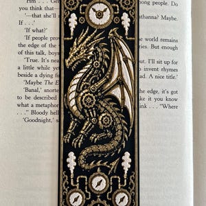 May include: A rectangular bookmark featuring a black background with a gold and white mechanical dragon design. The dragon is surrounded by gear motifs and four circular clock-like elements with white faces and gold hands, arranged at the top and bottom.