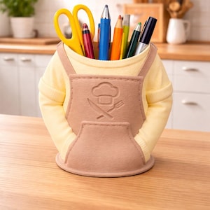 May include: A novelty desk organizer shaped like a chef's uniform, with a yellow shirt and a tan apron. The apron has a pocket and a chef's hat and utensils design. It holds pens, pencils, and scissors.