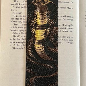 May include: A rectangular black bookmark featuring a detailed, raised relief illustration of a cobra snake. The snake is rendered in shades of metallic gold, bronze, and dark brown, with its hood flared and body coiled across the length of the bookmark.