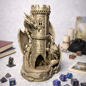 May include: A metallic gold-colored dice tower shaped like a stone castle turret with two sculpted dragons wrapped around the exterior. The tower features a crenelated top, a small arched window, and a large arched opening at the base for dice.