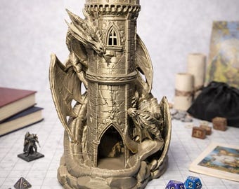 May include: A metallic gold-colored dice tower shaped like a stone castle turret with two sculpted dragons wrapped around the exterior. The tower features a crenelated top, a small arched window, and a large arched opening at the base for dice.