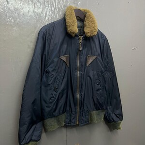 Puede incluir: Chaqueta bomber azul marino con cuello de borrego marr&oacute;n y detalles de cuero marr&oacute;n. La chaqueta tiene cremallera, dos bolsillos delanteros y pu&ntilde;os y dobladillo de canal&eacute; verdes. Colgada de una percha de madera.