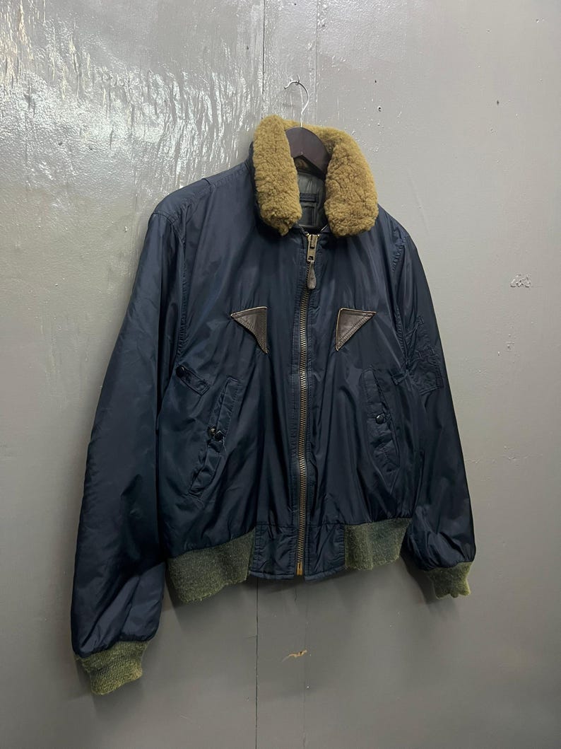 Puede incluir: Chaqueta bomber azul marino con cuello de borrego marr&oacute;n y detalles de cuero marr&oacute;n. La chaqueta tiene cremallera, dos bolsillos delanteros y pu&ntilde;os y dobladillo de canal&eacute; verdes. Colgada de una percha de madera.