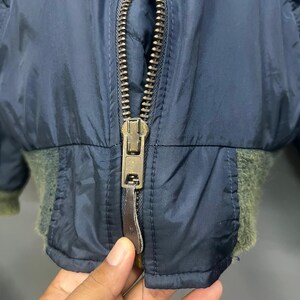Puede incluir: Primer plano de una chaqueta bomber azul marino con cremallera de lat&oacute;n y pu&ntilde;os de canal&eacute; verde oliva. La chaqueta tiene un forro acolchado y un tirador de cremallera de cuero. La cremallera est&aacute; parcialmente abierta.