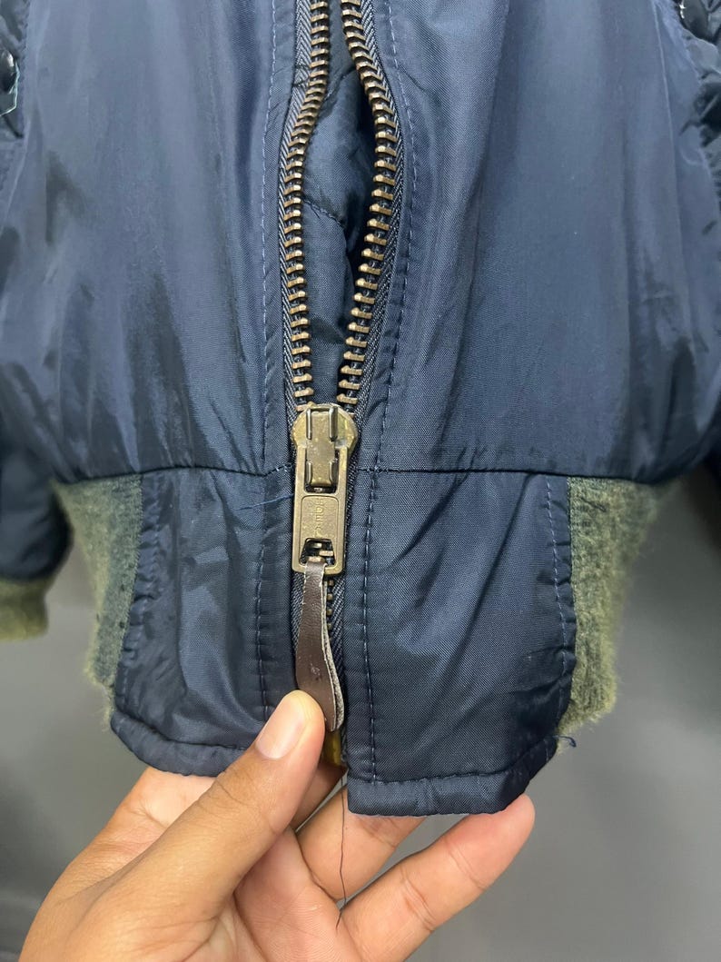 Puede incluir: Primer plano de una chaqueta bomber azul marino con cremallera de lat&oacute;n y pu&ntilde;os de canal&eacute; verde oliva. La chaqueta tiene un forro acolchado y un tirador de cremallera de cuero. La cremallera est&aacute; parcialmente abierta.