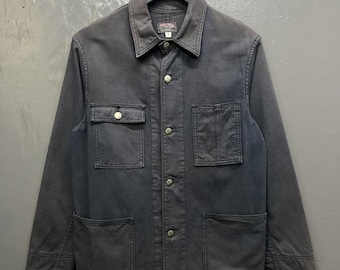 May include: Dark gray jacket with a button-up front, two chest pockets, and two lower pockets. The jacket has a collar and is hanging on a black hanger. The fabric appears to be a sturdy cotton or canvas.