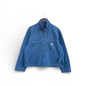 May include: A blue denim jacket with a zip-up front, two chest pockets, and two side pockets. The jacket has a small white tag on the right chest pocket and is hanging on a black hanger.