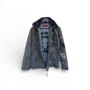 May include: A gray denim jacket with a black faux fur collar, adorned with various patches and a graphic tee visible underneath. The jacket has a zip closure and a drawstring at the waist. The patches include text and graphic designs.