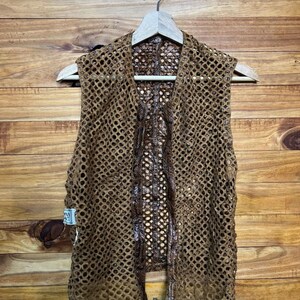 May include: A brown, open-weave vest with a unique, textured design. The vest is hanging on a wooden hanger against a wooden background. The vest has a distinctive pattern and appears to be handmade.