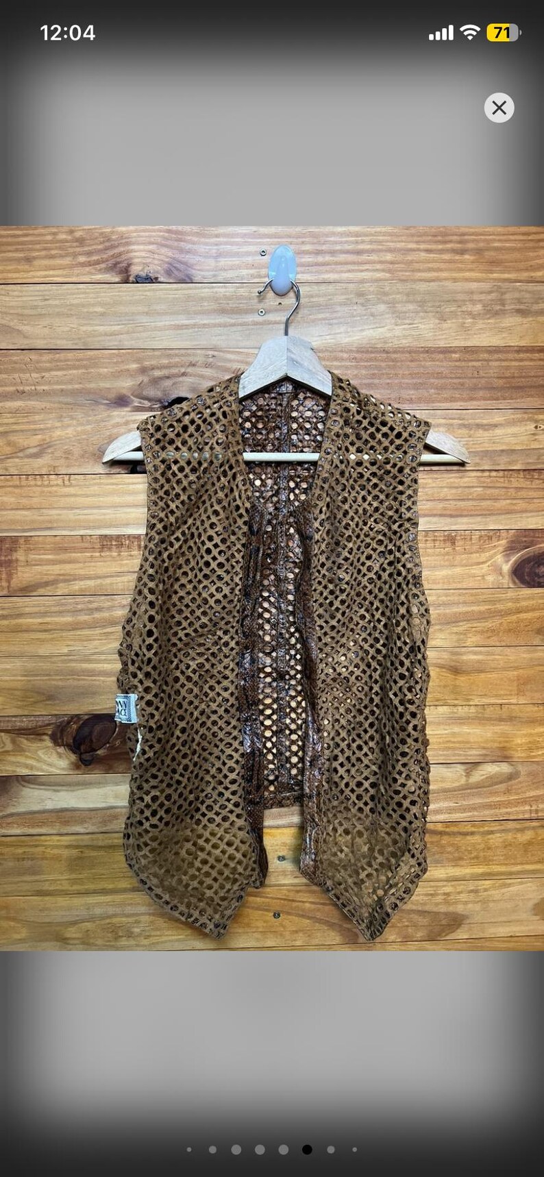 May include: A brown, open-weave vest with a unique, textured design. The vest is hanging on a wooden hanger against a wooden background. The vest has a distinctive pattern and appears to be handmade.