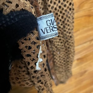 May include: Close-up of a brown, open-weave garment with a black accent. A tag with the text "GIA VERS" is attached. The garment appears to be made of a textured material, possibly leather or a similar fabric.