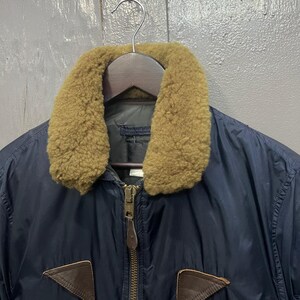 Puede incluir: Chaqueta bomber azul marino con cuello de borrego color canela y parches de cuero marr&oacute;n. La chaqueta tiene una cremallera de lat&oacute;n y est&aacute; colgada de una percha de madera. Una prenda de abrigo cl&aacute;sica.