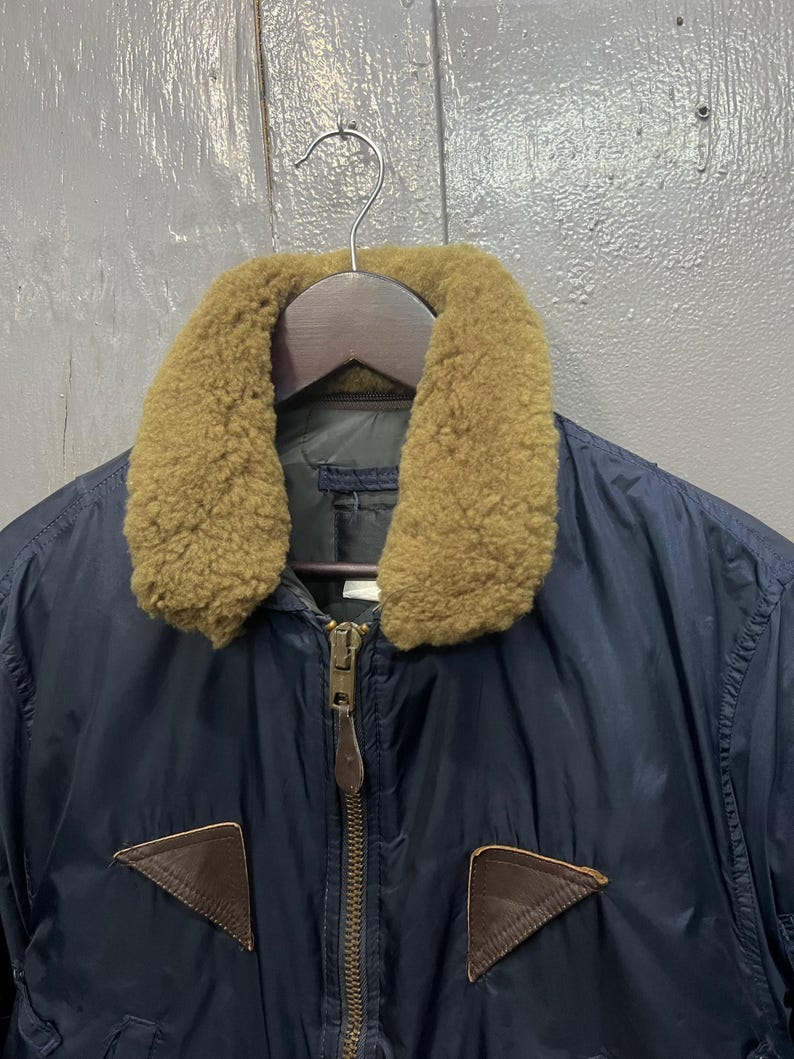 Puede incluir: Chaqueta bomber azul marino con cuello de borrego color canela y parches de cuero marr&oacute;n. La chaqueta tiene una cremallera de lat&oacute;n y est&aacute; colgada de una percha de madera. Una prenda de abrigo cl&aacute;sica.