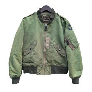 May include: Olive green bomber jacket with a zip-up front, two front pockets, and a patch on the upper left sleeve. The jacket has a ribbed collar, cuffs, and hem. The jacket is hanging on a black hanger.